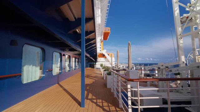 Modern cruise ship exterior deck featuring wooden walkway along blue hull with cabin windows. Outdoor seating area with chairs and tables under clear sky with ocean horizon visible.