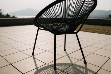 Obraz premium Modern black metal chair with string seat sits on tiled patio. Scenic view of mountains, sea in background. Outdoor furniture design for terrace garden relaxation. Minimalist patio decor with