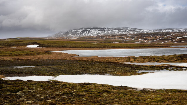 Tundra landscape showing patches of snow and a lake under a cloudy sky. Iceland Icelandic landscape
