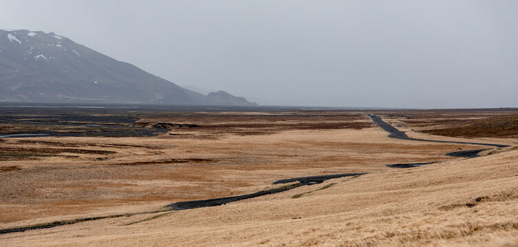 Panoramic landscape view of a desolate, valley in Iceland golden grass, dark volcanic terrain, winding road stretching into the distance, and a snow-dusted mountain under an overcast sky.