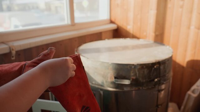White artisan prepping kiln with gloves, studio assistant checks kiln seal, apprentice adjusts apron, hands tuck gloves before firing, warm sunlight through window, wooden walls, clay tools