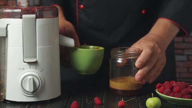 Chef prepares fruit puree by pouring blended mixture from green bowl into glass jar, with fresh raspberries and green apples arranged on wooden table in a cozy kitchen setting