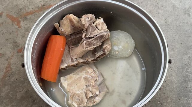 Boiled bones with carrot and onion in a metal pot of broth. Homemade soup base, bone stock, cooking ingredients, simple meal, kitchen food