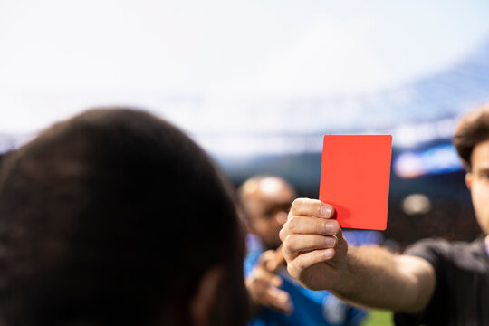 Football referee holds a red card while players protest the dismissal decision, creating controversy over unsportsmanlike conduct and a dramatic send-off during the soccer match on the stadium pitch.