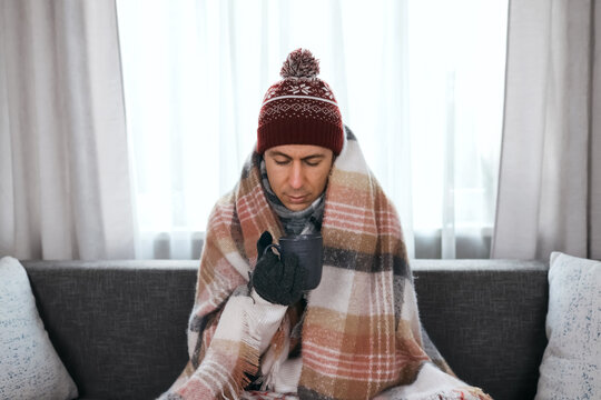 Freezing man wrapped in a blanket holds a warm mug at home. High heating costs and the energy crisis cause indoor cold weather, highlighting a severe home heating problem.
