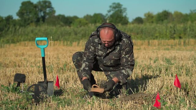 Male treasure hunter in camouflage clothing kneels in a field, examining a discovered object while surrounded by red flags marking the dig site and a metal detector and shovel nearby
