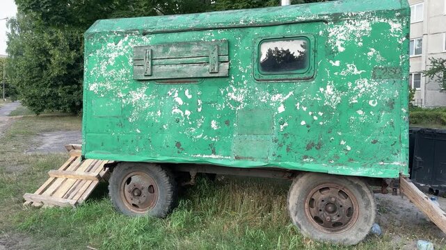 Old green trailer with peeling paint and rusty wheels parked on grass. Vintage caravan, weathered metal, rural life, abandoned transport