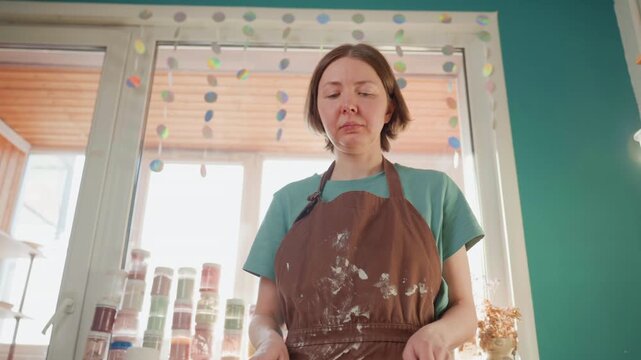 Focused white woman pottery instructor testing sponge tools near window, holding sponge on stick, rows of glaze jars behind, thoughtful expression, reaching for brush and container, calm instructional