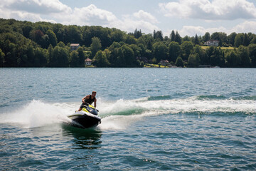 Fototapeta premium Young man rides fast jet ski on summer lake water. Male person makes sharp turn with motorboat creates big splash spray wave. Active water sport lifestyle on sunny day near green forest shore. Person