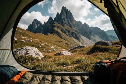 View from inside camping tent towards massive rocky mountain peaks under cloudy sky. Sleeping bag, backpack sit on ground near tent opening. Summer landscape shows grassy hills, large stones in