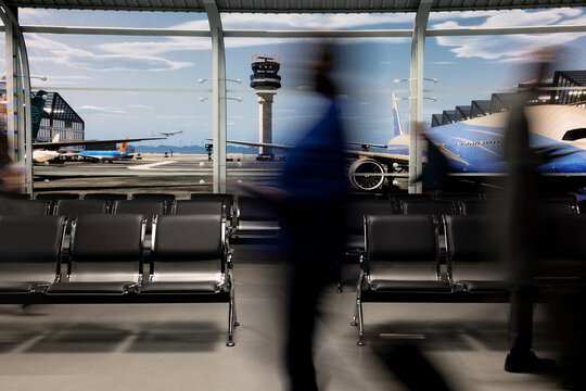Travelers move through a busy airport departure terminal in blurred motion amid the holiday rush, carrying luggage with the runway and control tower behind as passengers prepare to fly home.