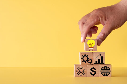 Hand placing a wooden block with a yellow lightbulb icon on top of a business icon pyramid. Concept of creative idea, strategic planning, financial growth, and global business success.