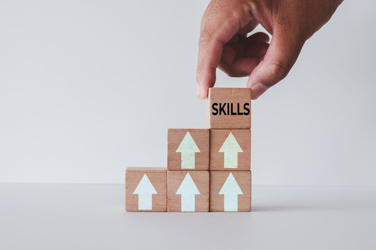 Hand placing a wooden block with the word SKILLS on top of a staircase made of blocks with upward arrows. Concept of personal development, career growth, upskilling, and continuous improvement.