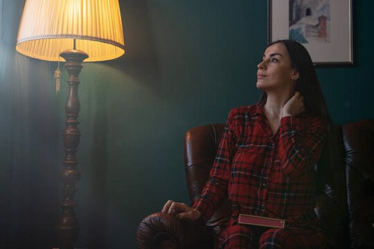 Thoughtful woman sitting with book beside glowing floor lamp