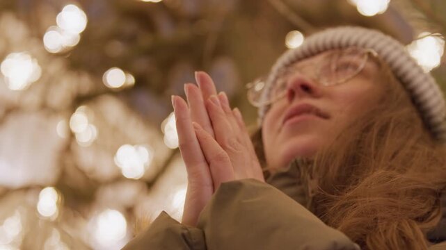 Caucasian woman under warm bokeh lights, glasses and knit beanie, parka and clasped hands near face, hopeful upward gaze, silent wish beside evergreen branches, golden glow and intimate closeup