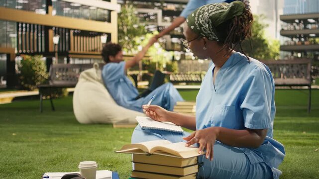 The medical student in blue scrubs reads and writes in a textbook during an outdoor study session, sitting on the grass with stacked books and classmates in the background, for science education.
