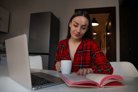 Woman reading a book beside laptop and mug at home