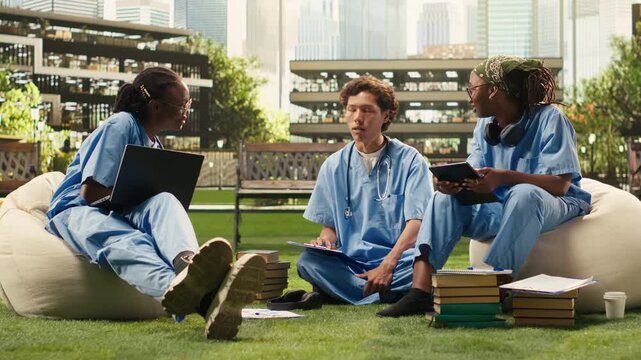 Medical students discuss coursework with a laptop and study materials, seated on beanbags beside stacked books and notes in a sunny campus courtyard during peer tutoring and outdoor group study.