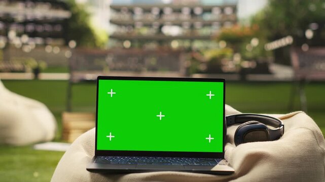 The laptop displays a green screen mockup on a modern campus beanbag with blurred buildings, designed for editable screen replacement in education content, representing university life.