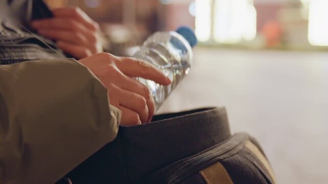 Hands holding water bottle against backpack. Gentle sip and cap twist, condensation on plastic, casual jacket sleeve, ambient bokeh of busy terminal, candid moment of refreshment and brief rest