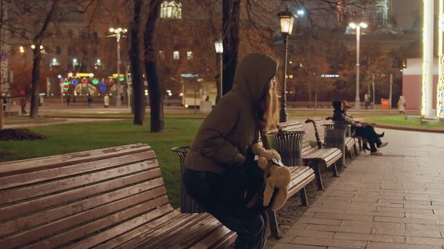 White woman sitting on park bench clutching teddy in puffer hoodie under warm streetlight glow, autumn leaves on wet pavement, quiet city square in evening, pensive mood, frames evoke student, late