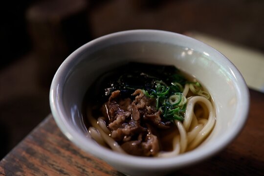 古い飲食店の木製テーブル。白碗に入った肉うどん。暗めの店内。