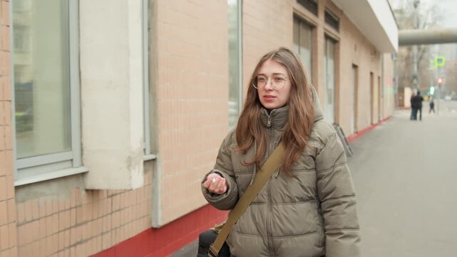 Young white woman strolling sidewalk beside brick building holding vape, pausing like commuter, adjusting hair like student, inhaling and exhaling vapor, glancing at storefront windows, neutral