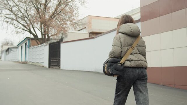 White woman walking urban alley carrying messenger bag puffer jacket, overcast sky, muted tones, quiet residential block, back view, candid stride, autumn leaves, solitary commute mood.