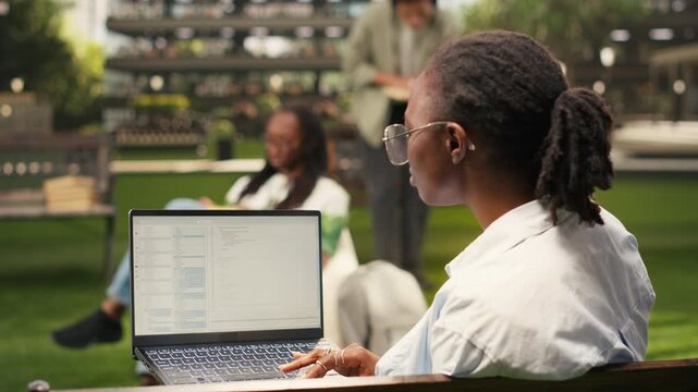 The laptop screen displays programming language in a terminal window as a student works in an outdoor campus study area, with classmates blurred in the background for focused coding practice.
