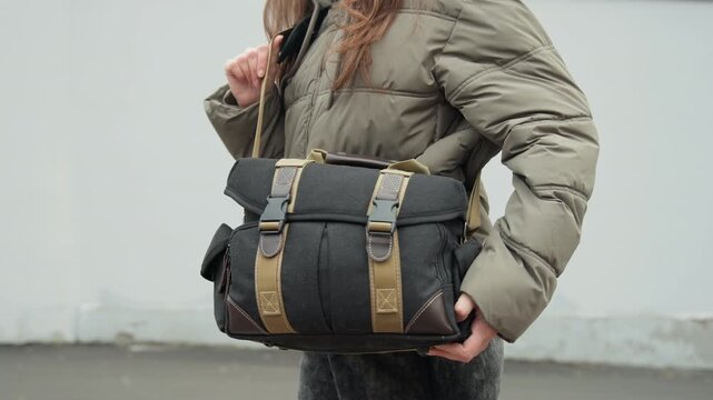 White woman closeup holding messenger bag detailed shearling texture, brass buckles, canvas panels, casual puffer jacket, tactile adjustment, urban street backdrop, product showcase feel.