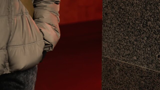 Individual paused near stone pillar at night. Person in padded coat reflected in urban night setting. Female silhouette in winter jacket pauses in moody city tunnel with reflections and shadows