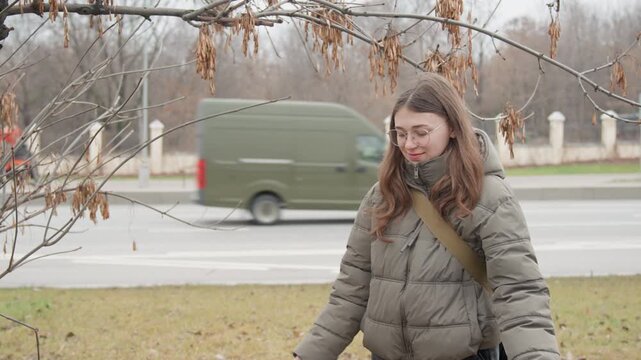 Wide shot woman releasing seeds from palms beside busy road, playful gesture and visible breath in cold air, puffer jacket and glasses set against moving cars and pale parkland, joyful candid moment