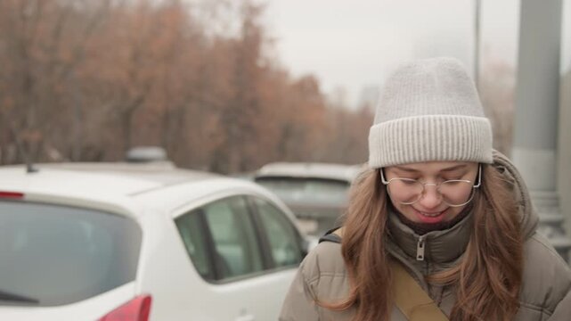 Young white woman outdoors looking down, wearing beanie and puffy coat on chilly city sidewalk, parked cars and lamppost in background, breath visible, pensive melancholic expression, commuter routine