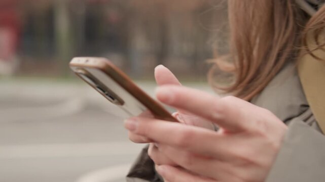 Closeup hands holding phone outdoors, urban commuter focused on messaging and scrolling through notifications while waiting by street with passing vehicles, neutral coat and phone case visible, soft