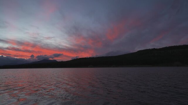 Dramatic sunset view over a tranquil lake, featuring vibrant red and purple clouds reflecting on the water, with silhouetted mountain ridges and forest in the distance.