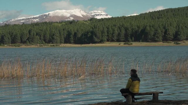 Man wearing a yellow jacket sitting on a wooden bench overlooking a calm lake with snow-capped mountain peaks in the background.