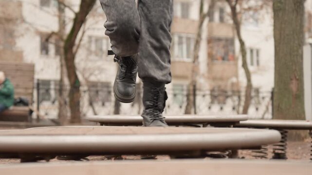 Person standing in park near spring platform, boots step onto circular disc as platform gives faint wobble, muted winter light, bare trees and distant apartment facades frame contemplative solo moment