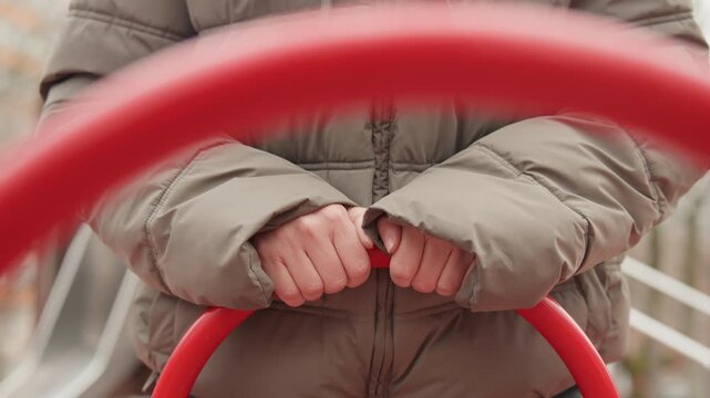 Closeup hands gripping red bar at empty playground, person in olive puffer jacket holding metal loop, muted winter light, shallow depth, soft bokeh, contemplative stillness, tactile knuckles