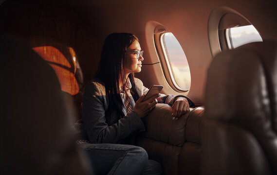 A dark haired woman, wearing glasses and a grey blazer, holds a smartphone and gazes quietly out the oval window of a private jet, her face lit by deep warm amber tones from outside.