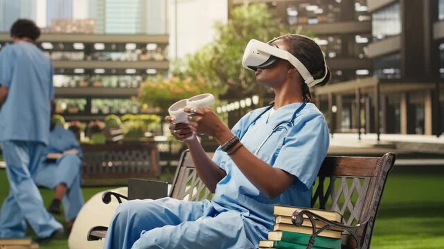 The woman medical student wears VR glasses and raises the controllers during medical training on campus, sitting on a bench in blue scrubs, using virtual reality for healthcare learning.