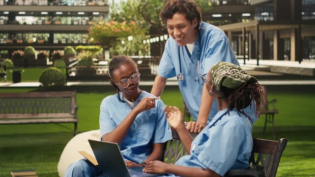 The group of peers in blue scrubs discusses coursework on a campus bench, using a laptop, open book and study materials for outdoor learning, student collaboration and medical training.