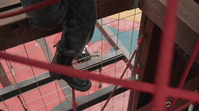 Boots on high steel frame worker navigates red rope net above city, worn black boots step across rusted beam, tense balance and careful foothold, metal grid and safety gear glimpsed, vertical