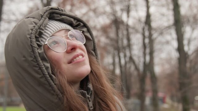 White woman in park gazing upward with quiet confidence, hooded parka and glasses soft smile, autumn trees and playground background, layered winter fashion and relaxed posture, candid portrait