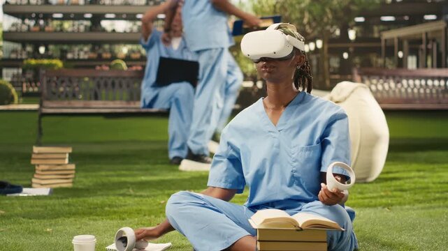 The Black woman medical student wears a VR headset and raises the controllers during medical training, dressed in blue scrubs, using virtual reality for healthcare learning on campus.