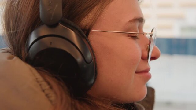 Caucasian student wearing headphones on commute, side profile closeup adjusting black over ear headphones, round glasses, brown coat, bus window city blur, soft winter light, serene smile, ambient