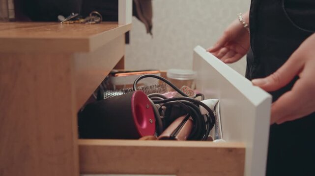 White woman grabbing pink straightener from drawer, hallway closet with coat and keys visible, wooden console holding hairdryer and accessories, close crop on midriff and bracelet, candid morning prep