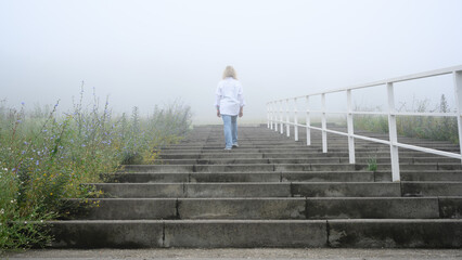 Obraz premium Woman walking up concrete stairs into dense fog. Solitary person in white shirt and jeans ascending outdoor steps towards the misty unknown