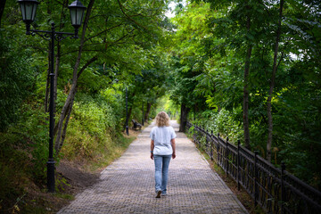 Obraz premium Rear view of a blonde woman walking on a park path. Female pedestrian strolling along a paved walkway surrounded by green trees. Outdoor summer leisure and active lifestyle
