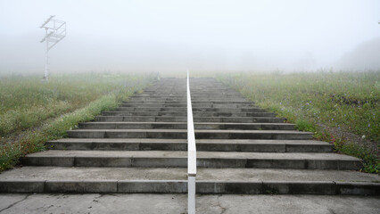 Fototapeta premium Concrete stairs ascending into dense fog. Empty outdoor steps with white handrail leading to the unknown, old stairway in mist. Copy space for text
