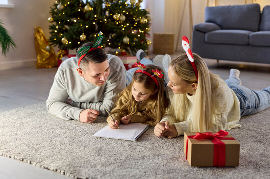 Family child writing Christmas letter near tree. Parents lie beside on carpet, guiding a wish list for Santa amid winter holiday lights, gifts, and cozy decor. Warm festive family bonding at home.